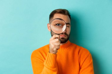 Komik adam büyüteçten bakıyor, bir şeyi arıyor ya da araştırıyor turkuaz arka planda turuncu süveter giyiyor.