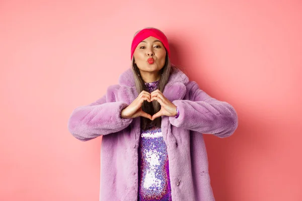 Romantika a Valentýn. Happy asian senior woman showing heart sign, I love you gesture, pucker lips for kiss, standing over pink background — Stock fotografie