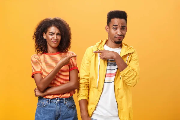 Friends getting shocked someone thought they are dating. Displeased mocking african american female frowning and grimacing from aversion while pointing at guy who indicating at girl over orange wall — Stock Photo, Image