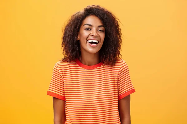Waist-up πλάνο του εξερχόμενου ευτυχισμένη γοητευτική μελαχρινή γυναίκα με σγουρά μαλλιά γέλιο χαρούμενα ποζάρουν σε ριγέ μοντέρνα t-shirt πάνω από πορτοκαλί φόντο απολαμβάνοντας ωραία casual συνομιλία — Φωτογραφία Αρχείου