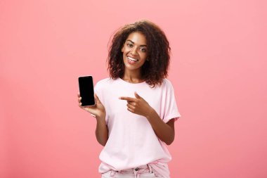 Bu telefon modelini öneriyorum. Güzel, Afro-Amerikan kıvırcık saçlı, şık elbiseli, elinde akıllı bir kadın ve pembe duvarın üzerinde havalı bir uygulama önerirken cihaz ekranını gösteren mutlu bir afro-amerikan kadını.