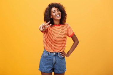 Dost canlısı kaygısız ve mutlu görünümlü uzun boylu Afro-Amerikan kadın modeli, kıvırcık saç stili olan, kalça eğen kafasının üstünde el ele tutuşan ve turuncu duvarın üzerinden neşeyle gülümseyen.