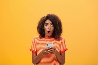 Şok olmuş ve endişeli afro saç stili olan afro saçlı bir kadının portresi. Endişeden gözleri patlıyor ve elinde akıllı telefon ile panikleyip korkunç bilgiler veriyor.