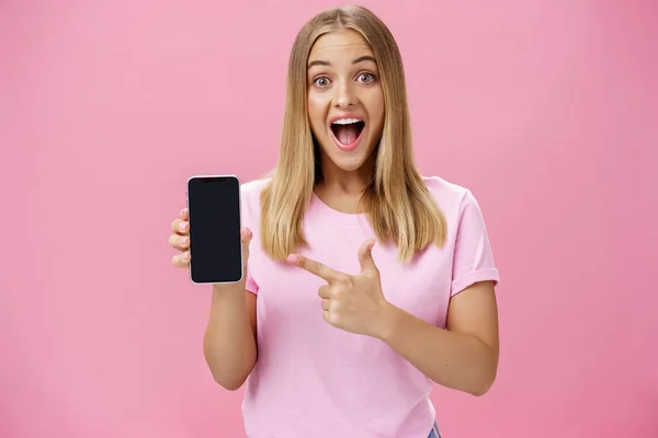 Portrait of excited and amused charming pleased female client using new smartphone advicing friends to buy awesome gadget holding cellphone and pointing at device screen with broad amused smile — Stock Photo, Image