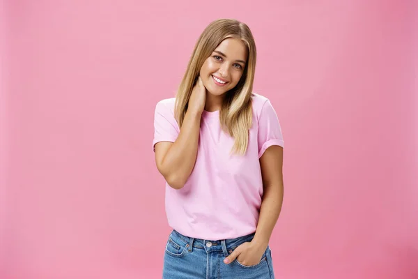 Portrait of shy and timid attractive european woman with fair hair in casual outfit touching neck silly tilting head and smiling cute at camera standing over pink background — Stock Photo, Image
