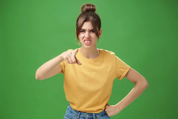 Portret van arrogante, onbeleefde jonge vrouw in geel t-shirt die wijst naar camera 's die kraken en griezelen van minachting en minachting die staan te oordelen tegen een groene achtergrond — Stockfoto