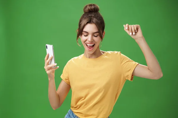 Cool and stylish happy woman with tattoo and gap teeth in yellow t-shirt dancing joyfully listening music in wireless earphones holding smartphone closing eyes and smiling from delight over green wall — Stock Photo, Image