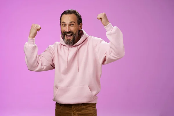 Ja, wen interessiert schon das Alter. Sorglos entzückte glückliche alte Mann bärtig in trendigen rosa Kapuzenpullover heben die Fäuste freudig triumphierend Spaß freudig Sieg feiern Erfolg erreichen Ziel, posiert lila Hintergrund — Stockfoto