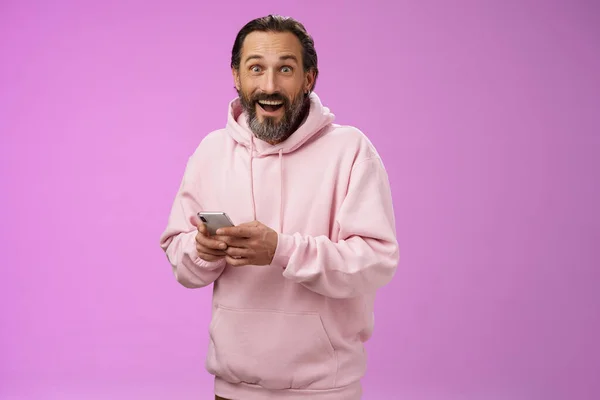 Amüsiert glücklich fasziniert erwachsenen erwachsenen kaukasischen bärtigen Mann in rosa Kapuzenpullover keuchend beeindruckt halten Smartphone-Look Kamera erstaunt Kauf cool neues Handy, erstaunt, wie bequem — Stockfoto