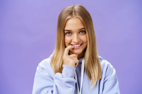 Close-up di femminile e tenera ragazza con i capelli biondi e la pelle abbronzata mordere dito flirty e timido sorridente carino a macchina fotografica in piedi in moda over-size felpa con cappuccio, flirtare su sfondo viola — Foto Stock