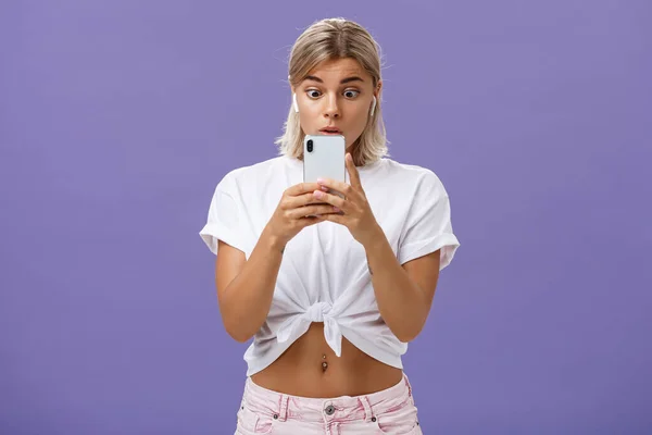 Menina assistindo vídeo emocionante nas mídias sociais em fones de ouvido sem fio segurando smartphone perto da cara estourando os olhos na tela do smartphone olhando para o gadget com interesse e curiosidade sobre o fundo roxo — Fotografia de Stock