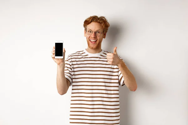 Concepto de tecnología y comercio electrónico. Joven con el pelo rojo mostrando pulgares hacia arriba y la pantalla del teléfono inteligente en blanco, recomendando aplicación, fondo blanco — Foto de Stock