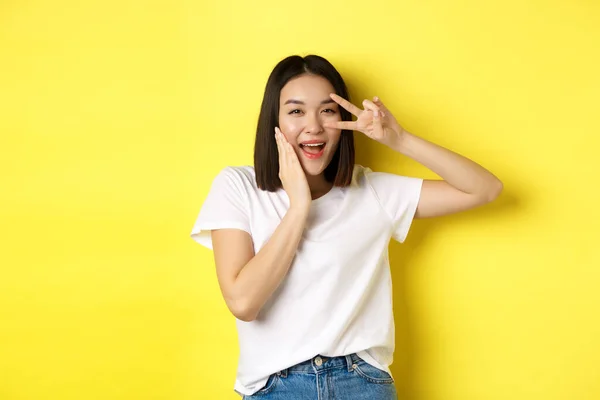 Schöne asiatische Mädchen in weißem T-Shirt posiert mit der Hand auf der Wange, zeigt Peace Zeichen auf Auge, steht über gelbem Hintergrund — Stockfoto