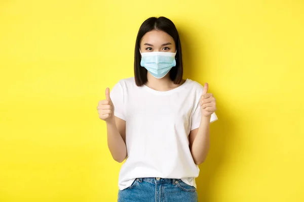 Covid-19, karantän och socialt distanserande koncept. Glada asiatiska kvinna i medicinsk mask och vit t-shirt visar tummen upp i godkännande, beröm bra affär, gul bakgrund — Stockfoto