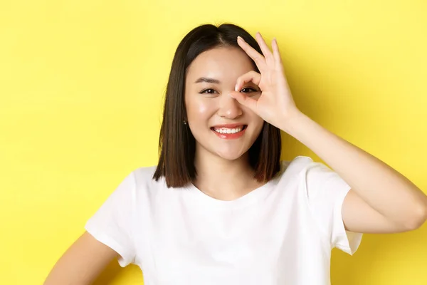 Lähikuva söpö aasialainen tyttö tunne onnellinen, osoittaa OK merkki silmään ja hymyilee, seisoo yli keltainen tausta — kuvapankkivalokuva