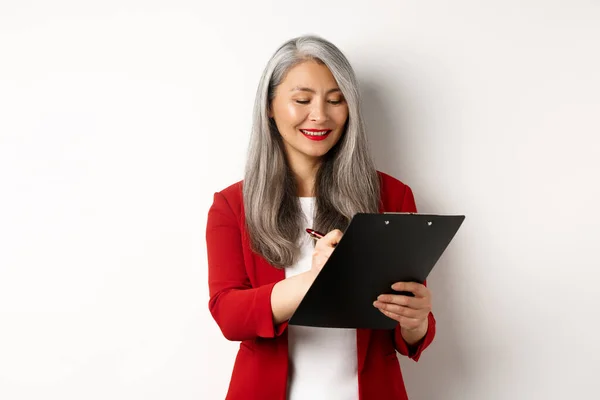 Succès asiatique senior femme d'affaires avec cheveux gris prendre des notes sur presse-papiers, inspecter l'entreprise, debout sur fond blanc — Photo