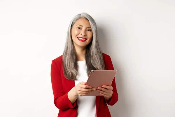 Unternehmen. Erfolgreiche Seniorin arbeitet mit digitalem Tablet und lächelt, steht im roten Blazer vor weißem Hintergrund — Stockfoto