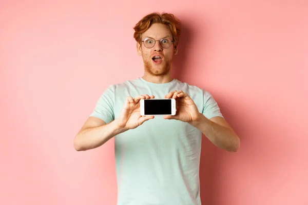 惊讶的红头发男人喘息着，敬畏地盯着相机，横向地展示着空白的智能手机屏幕，站在粉色的背景上 — 图库照片