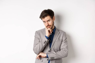 Kararsız iş adamı karar veriyor, endişeli yüzüyle kameraya dalgın dalgın bakıyor, şüpheleri var, beyaz arka planda duruyor.