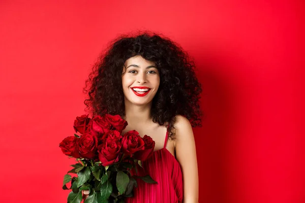 Modello femminile alla moda felice con mazzo di rose rosse, sorridente e guardando allegro a macchina fotografica, sfondo di studio — Foto Stock