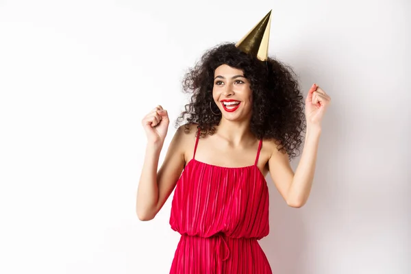 幸せな誕生日の女の子は、パーティーの帽子と赤いドレスを着て、踊り、楽しんで、白い背景を背景に立って祝う — ストック写真