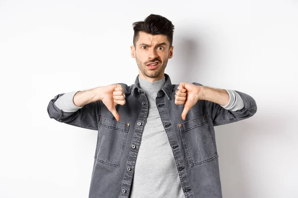 Teleurgesteld man toont duimen naar beneden en fronsen, kijkt sceptisch naar iets slechts, afkeer en afkeuring verschrikkelijk ding, staan op witte achtergrond — Stockfoto