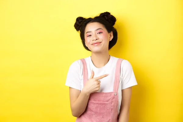 Elegante asiático adolescente menina com maquiagem romântica, sorrindo bonito e apontando o dedo para a direita no logotipo, de pé contra fundo amarelo — Fotografia de Stock