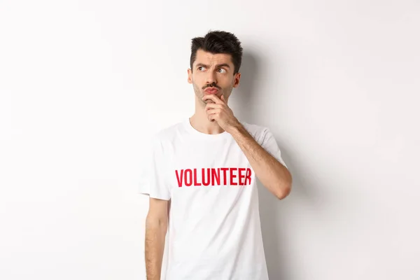 Varón joven reflexivo voluntario en soluciones de búsqueda de camisetas blancas, mirando la esquina superior izquierda y pensando, de pie sobre el fondo del estudio —  Fotos de Stock