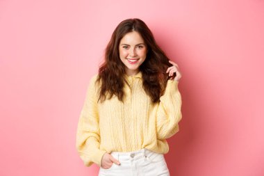 Güzel kız saçlarıyla oynuyor, gülümsüyor ve kameraya kendinden emin bir şekilde bakıyor, cazibe pozu veriyor, pembe arka planda duruyor.