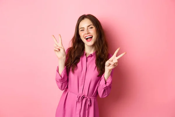 Θετική όμορφη κοπέλα σε κομψό φόρεμα, δείχνει v-υπογράψει την ειρήνη και χαμογελώντας ευτυχισμένη, θέτοντας κοντά στο ροζ φόντο — Φωτογραφία Αρχείου