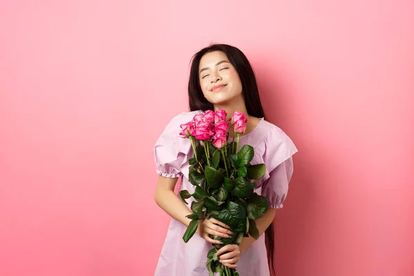 Sonhador asiático adolescente sentindo romântico, segurando flores e sonhando, imagem dia dos namorados data, vestindo vestido bonito, de pé sobre fundo rosa — Fotografia de Stock