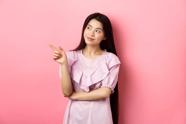 Ονειρεμένη Ασιάτισσα με φόρεμα, κοιτάζει και δείχνει αριστερά στο λογότυπο, ατενίζει τον σχεδιαστή, στέκεται απέναντι σε ροζ φόντο — Φωτογραφία Αρχείου