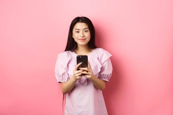 Online αγορές. Χαριτωμένο κορίτσι της Ασίας χαμογελά, κρατώντας το κινητό τηλέφωνο με χαρούμενο πρόσωπο, στέκεται στο φόρεμα σε ροζ φόντο — Φωτογραφία Αρχείου