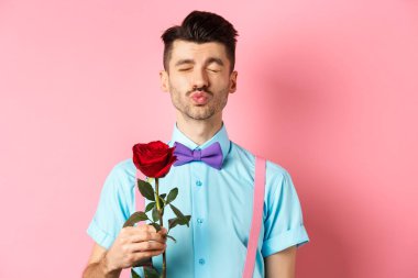 Sevgililer Günü 'nde sevgilisinden öpücük bekleyen sevimli ve komik bir adam. Kız arkadaşı için güzel kırmızı bir gül tutuyor. Pembe arka planda duruyor.
