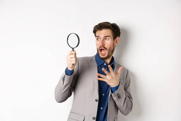 Chockad kille titta genom förförisk glas med häpen ansikte, kippar och hålla handen på bröstet rädd, står på vit bakgrund — Stockfoto