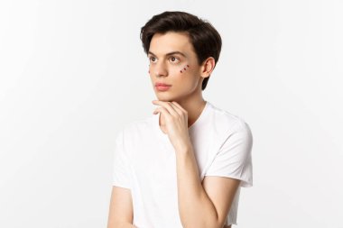 Millet, lgbtq ve güzellik konsepti. Güzel çift cinsiyetli adam, gözlerinin altında pırıltı, sol üst köşeye bakıyor, beyaz arka plana karşı poz veriyor.