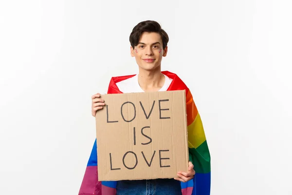 Glimlachende gay man activist holding teken liefde is liefde voor lgbt trots parade, het dragen van regenboog vlag, staande over witte achtergrond — Stockfoto
