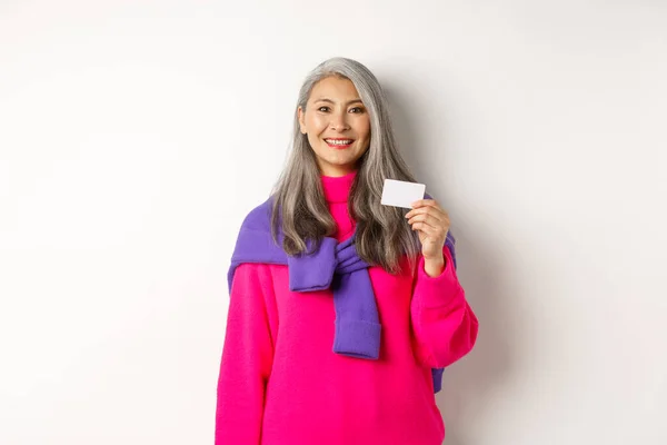 Concepto de compras. Sonriente asiático de mediana edad mujer con pelo gris mostrando tarjeta de crédito de plástico y mirando feliz, de pie sobre fondo blanco — Foto de Stock