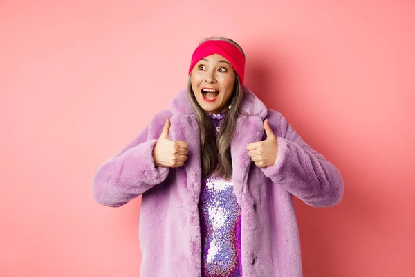 Mode- und Einkaufskonzept. Glückliche asiatische Seniorin in stylischer Kleidung schaut sich die tolle Werbung an, schaut in die linke obere Ecke und zeigt zustimmend Daumen nach oben — Stockfoto