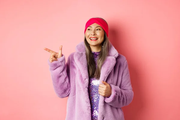 Glückliche ältere asiatische Frau lächelt, zeigt mit dem Finger nach links und sieht zufrieden aus, schaut sich Sonderaktionen an, rosa Hintergrund — Stockfoto