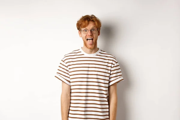 Vtipný mladý muž s rozcuchanými zrzavými vlasy a brýlemi ukazující jazyk, zírající do kamery, stojící nad bílým pozadím — Stock fotografie