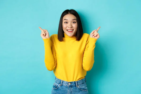 Concept beauté et mode. gai asiatique fille montrant deux promo offres pointant doigt sur gauche et droite annonces et souriant, debout sur fond bleu — Photo
