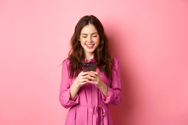 Ung kvinna shopping, chatta på mobiltelefon, leende när du läser smartphone skärm, står mot rosa bakgrund — Stockfoto