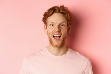 Şaşkın kızıl saçlı, tişörtlü, ağzı açık, kameraya hayran ve mutlu bakan, pembe arka planda duran erkek modelin vesikalık fotoğrafı.