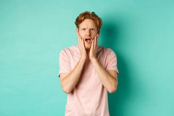 Geschokt en bezorgd jonge man met rood haar, staren naar camera bezorgd en ontroerende gezicht, staan in t-shirt tegen turquoise achtergrond — Stockfoto