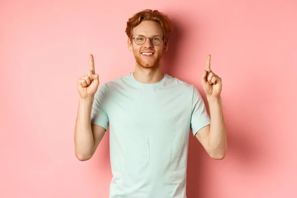 Atractivo joven en gafas y camiseta mostrando oferta promocional, señalando con los dedos hacia arriba y sonriendo a la cámara, de pie sobre el fondo rosa — Foto de Stock