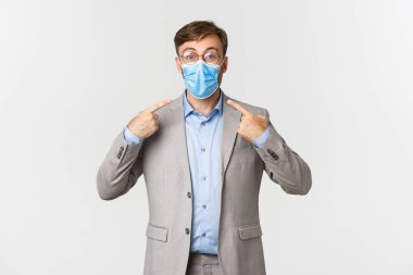 İş anlayışı, covid-19 ve sosyal mesafeler. Gri takım elbiseli ve gözlüklü heyecanlı iş adamının portresi, beyaz arka planda duran tıbbi maskeyi işaret ediyor.
