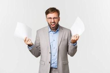 Gri takım elbiseli ve gözlüklü kızgın işadamının portresi, kötü rapordan hayal kırıklığına uğramış, elinde belgeler ve azarlayan bir çalışan, beyaz arka planda duruyor.