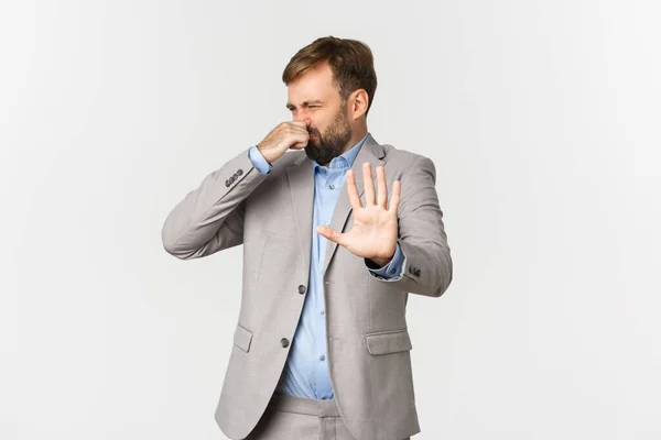 Porträt eines angeekelten bärtigen Geschäftsmannes im grauen Anzug, der etwas mit furchtbarem Geruch ablehnt, die Nase zuhält und ein Zeichen des Niedergangs setzt, vor weißem Hintergrund stehend — Stockfoto