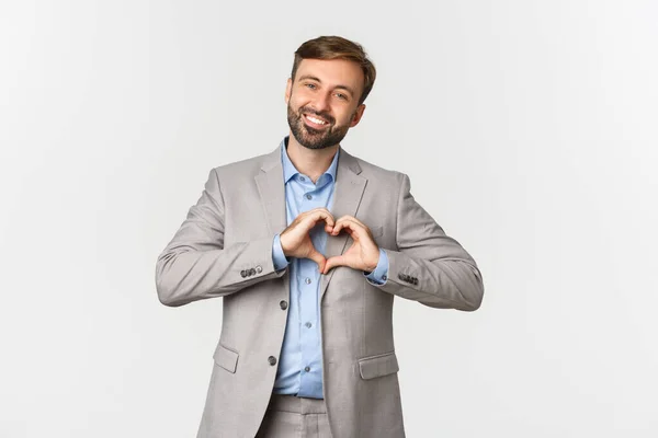 Retrato de belo homem de barba sorridente em terno de negócios, mostrando sinal de coração, expressar amor ou simpatia, de pé sobre fundo branco — Fotografia de Stock